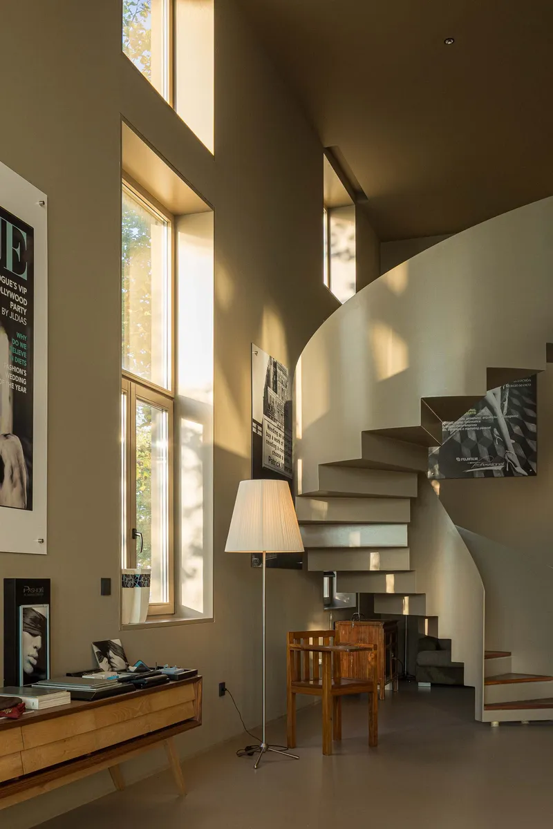 Curved floating staircase bathed in sunlight from tall windows beside framed posters