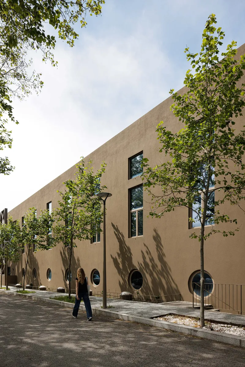 Street-facing facade with circular and rectangular windows and young trees casting shadows on beige stucco