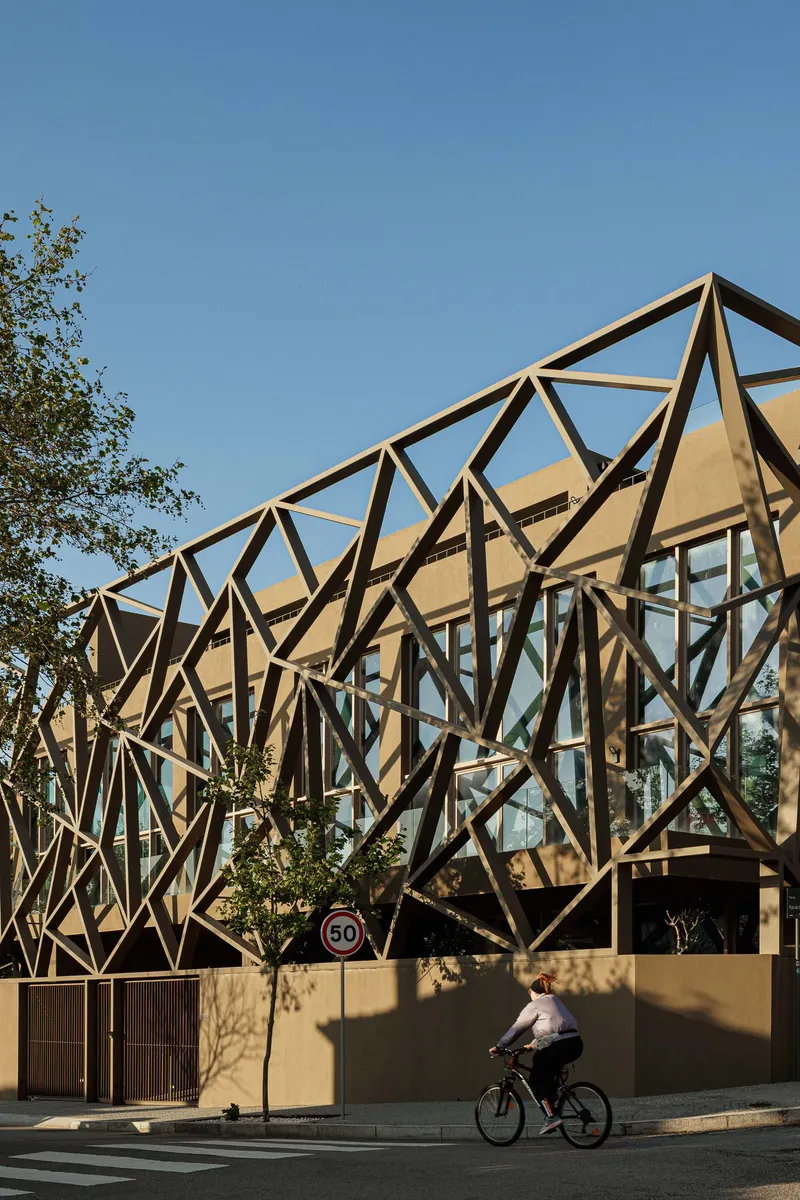 Diagonal steel lattice wrapping the upper volume as a cyclist passes beneath a clear blue sky