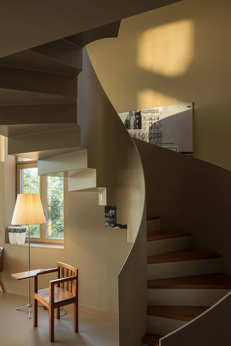 Spiraling stairwell with cantilevered treads and white plaster walls illuminated by afternoon light