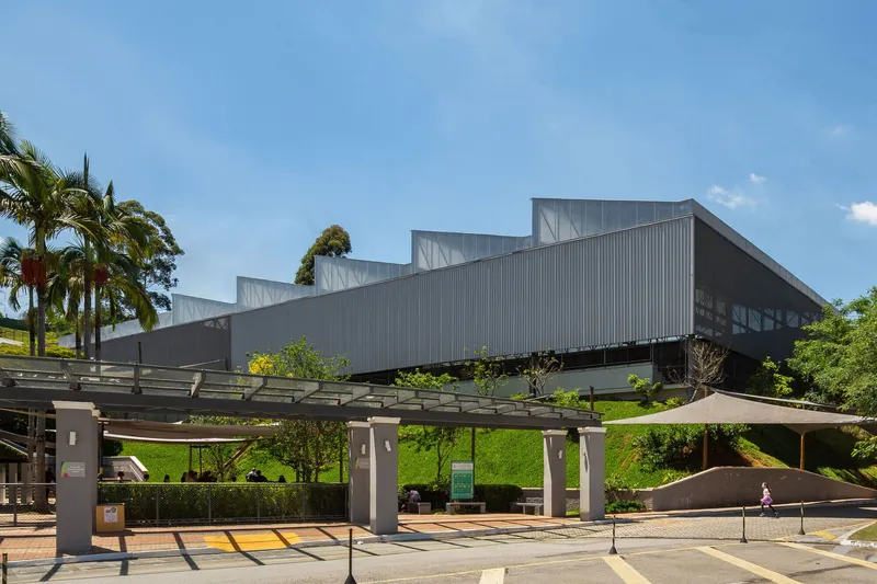Corrugated metal facade with stepped roofline rising above a pergola and grassy slope with palm trees