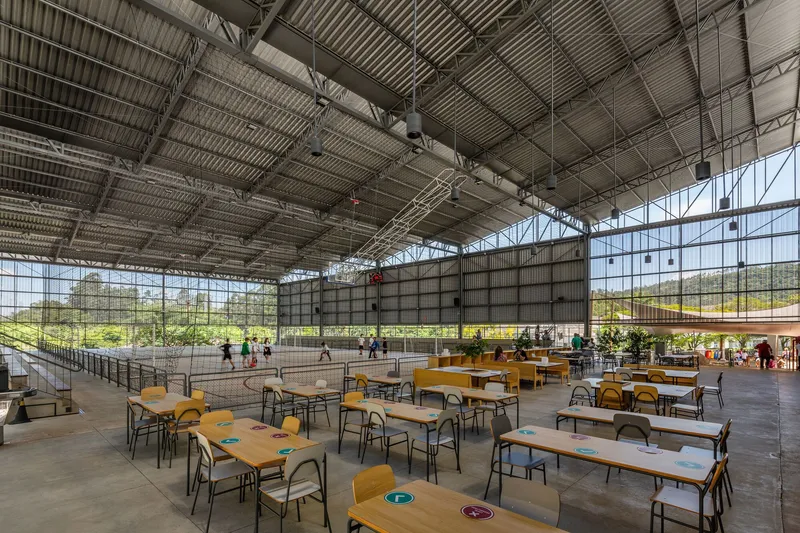 Dining area with wooden tables and benches beneath corrugated roof and curtain wall overlooking playing fields