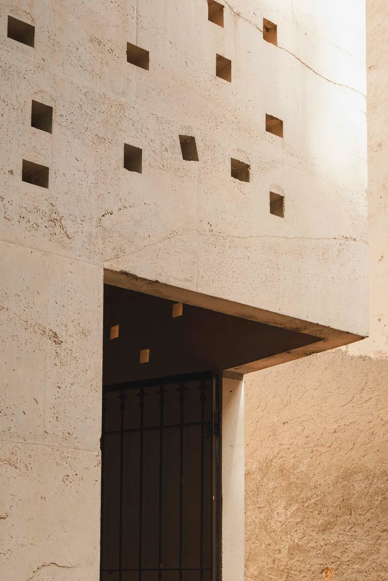Triangular canopy over a timber door with square openings dotting the concrete wall above