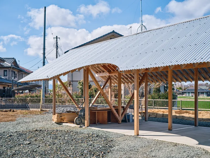 Open pavilion with exposed timber frame and corrugated metal roof sheltering a courtyard on a clear afternoon