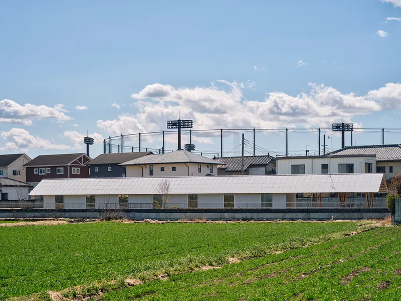 Low horizontal volume with white corrugated metal roof beyond green agricultural field and suburban houses