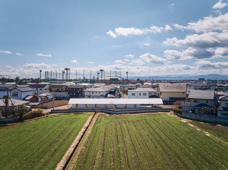 Aerial view of long horizontal building with white roof nestled between residential neighborhood and cultivated field