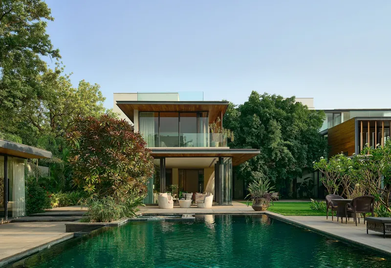 Two-storey glass and timber residence with pool terrace framed by mature trees under afternoon light