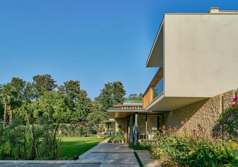 Cantilevered white stucco volume above stone-clad ground floor and paved entry path with planted beds