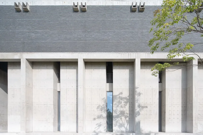 Facade with recessed concrete columns below grey brick wall and dappled tree shadows in daylight