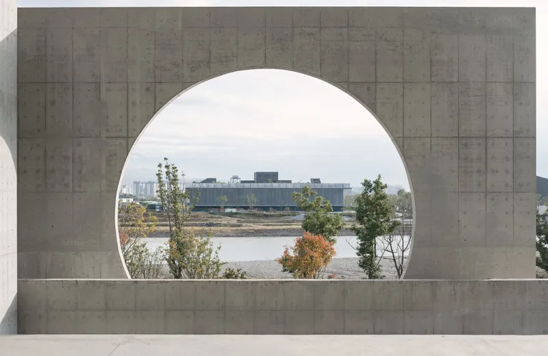 Large circular aperture in a board-formed concrete wall framing a view of the river and distant trees