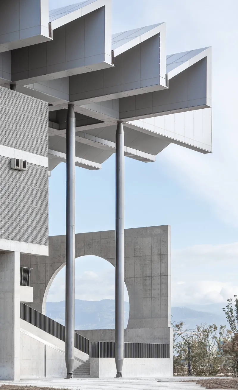 Steel columns supporting stacked concrete beams and sawtooth canopy against a cloudy mountain backdrop