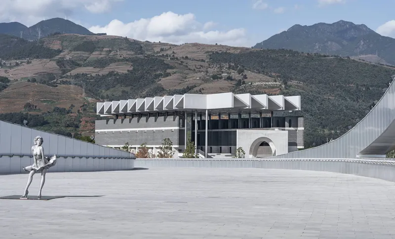 Plaza view with sculptural figure and sawtooth-roofed volumes set against rugged mountain terrain