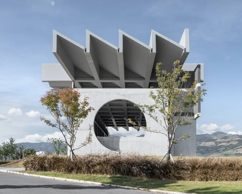White concrete facade with four folded roof planes above a large circular opening flanked by young trees