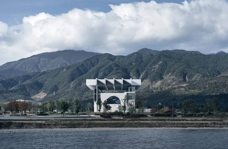Long view of the sawtooth roof structure across a waterfront with mountain range under cloudy skies