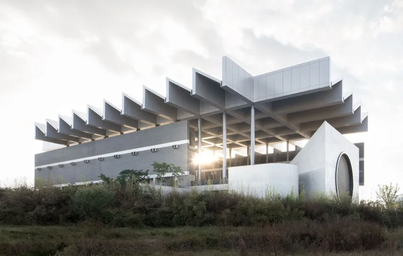 Sawtooth concrete roof structure with exposed columns backlit by sunset glow through vegetation