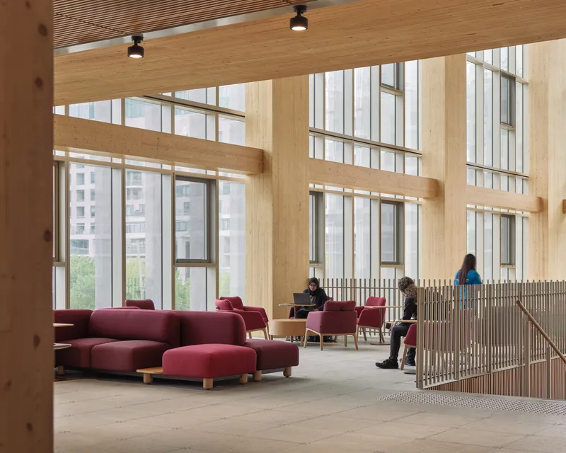Upper-level lounge with burgundy modular seating, wood slat railing, and tall windows framing buildings beyond