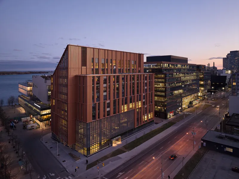 Aerial view of the copper-clad facade with vertical fins illuminated at dusk beside a waterfront boulevard