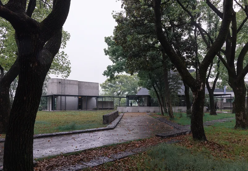 Paved path through mature trees leading to a concrete volume and glass-walled pavilions under overcast skies