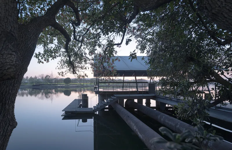 Elevated pavilion with metal roof and timber deck extending over still water at dusk beneath overhanging tree branches