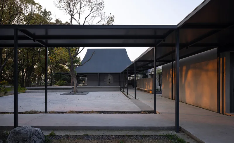 Covered walkway with steel frame and concrete floor connecting two pavilions with pitched roofs in early evening light