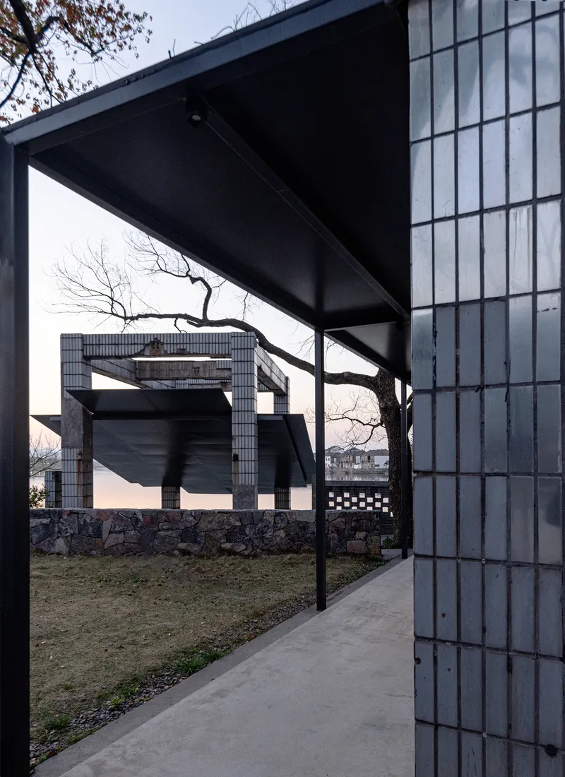 Concrete walkway beneath steel canopy framed by glass block wall and bare tree branches at dusk