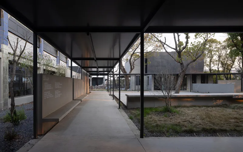 Corridor with steel columns and translucent roof panels opening onto courtyard with planted gravel and scattered trees