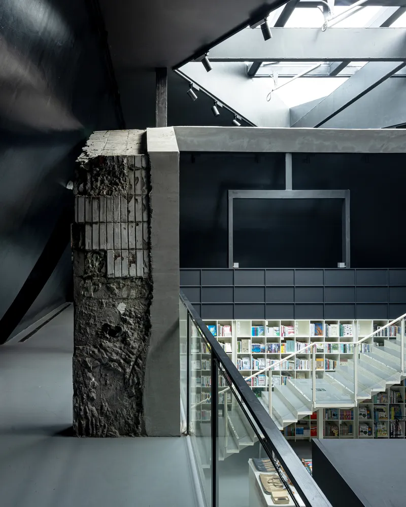Interior view of exposed concrete column with rebar detail beside glass-walled stairwell and bookshelves below skylight