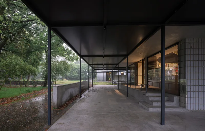 Covered entry walkway with slender steel columns and full-height glazing overlooking a park in wet weather