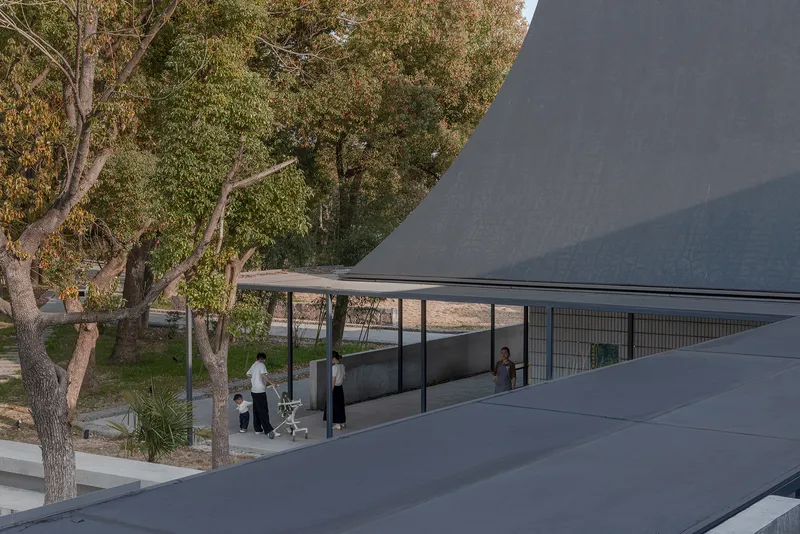 Curved gray roofscape framing a glass pavilion sheltered beneath mature trees with visitors below