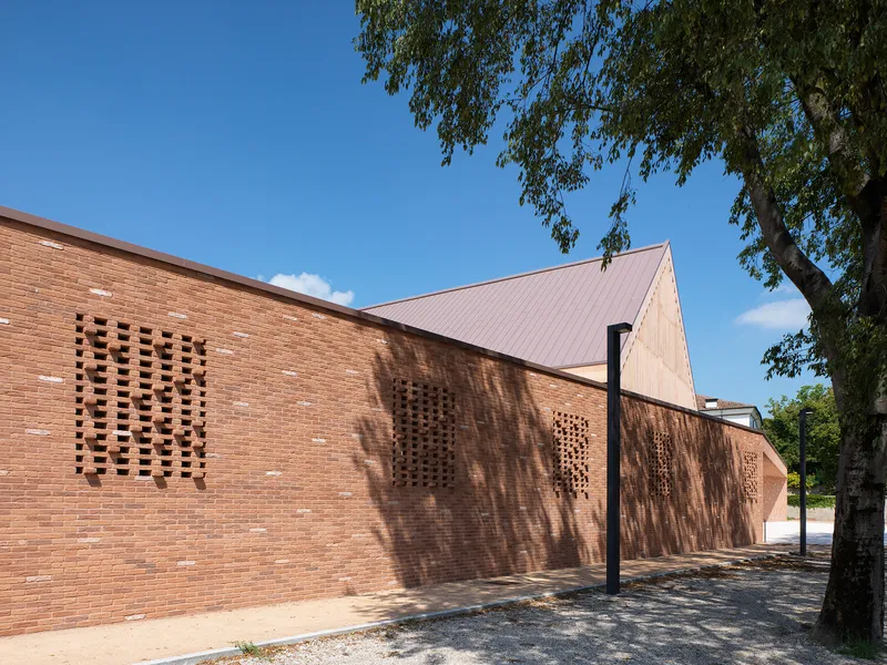 Perforated brick facade with angular gable roof under a clear blue sky and tree shadow