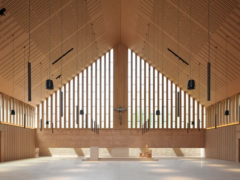 Altar wall with vertical timber screens, crucifix and suspended lighting beneath a pitched wood ceiling
