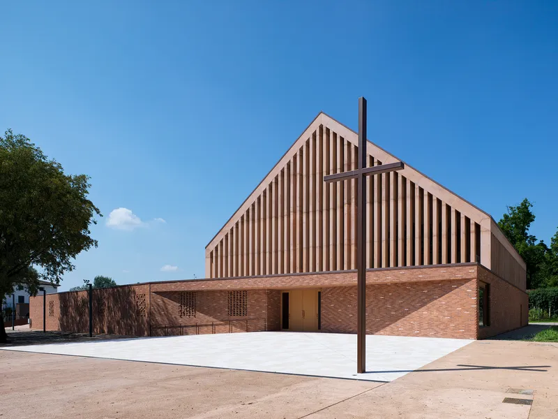 Gabled volume with vertical timber louvers above a brick base and a steel cross in the forecourt