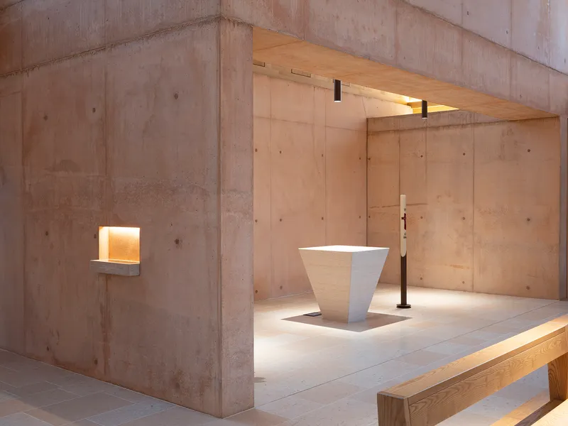 White stone baptismal font on a raised platform beneath exposed concrete beams and recessed wall niches