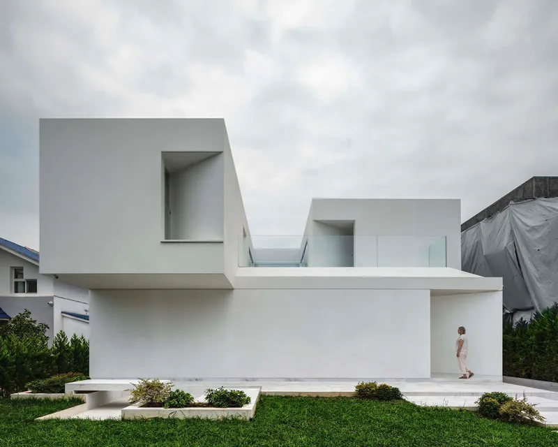 White rendered facade with cantilevered volumes and recessed entrance where a figure stands under overcast skies