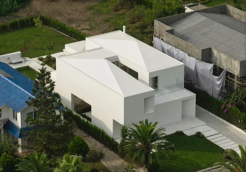 Drone view showing the folded roof planes above interlocking white volumes surrounded by palm trees