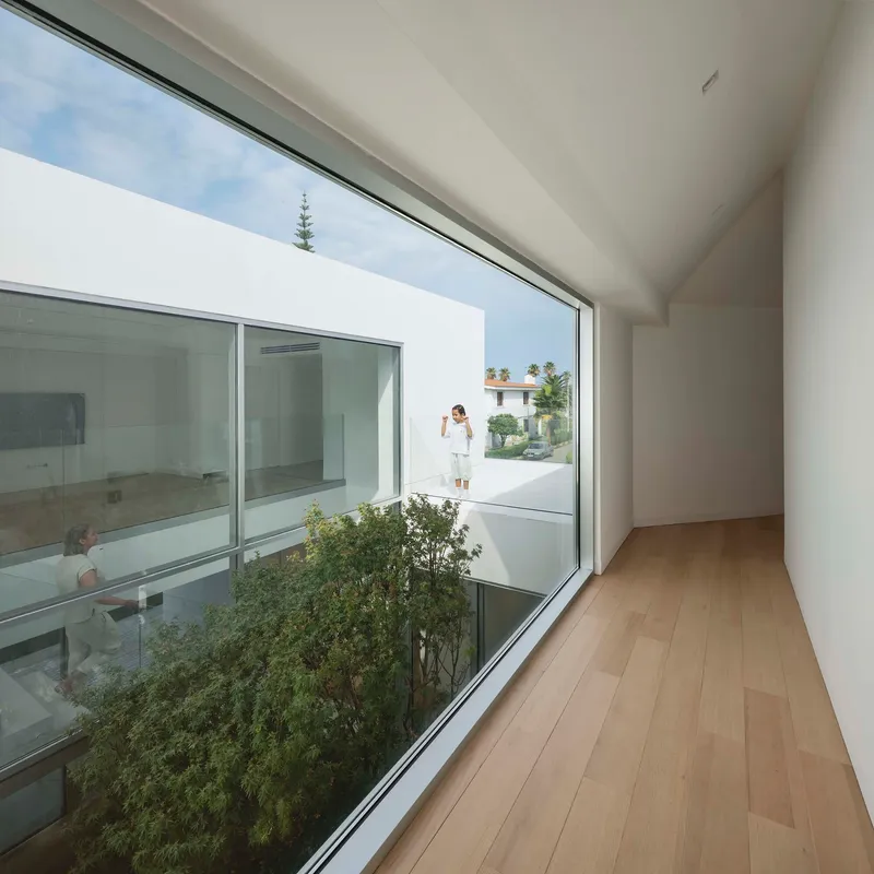 Timber-floored corridor with floor-to-ceiling glazing overlooking the planted courtyard and figure on exterior terrace