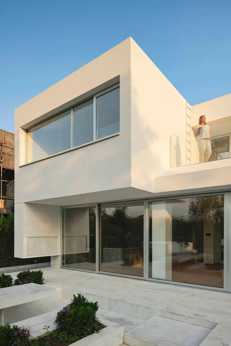 White rendered facade with full-height glazing and upper-level balcony at dusk with a figure visible