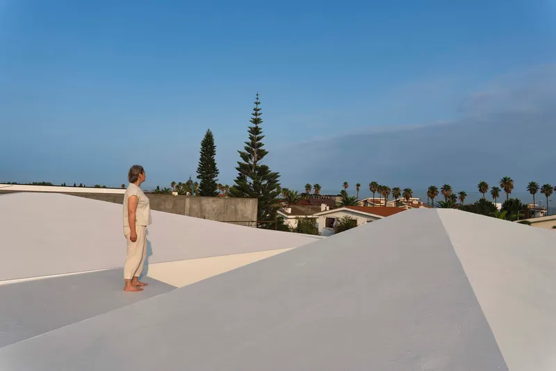 Rooftop terrace with white rendered volumes and a figure overlooking palm trees in the distance