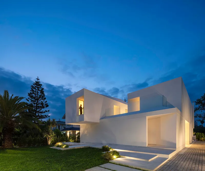 White cubic volumes with illuminated openings and recessed glass at twilight beside a lawn