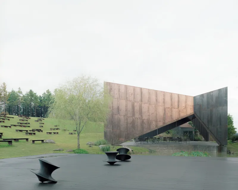 Weathered steel facade with sculptural metal benches on a wet plaza in spring