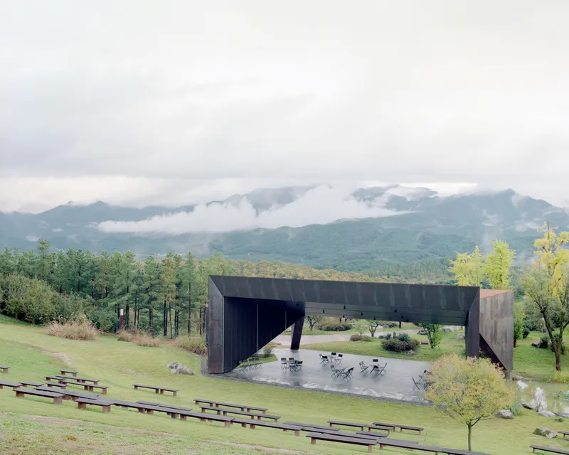 Cantilevered steel volume sheltering an outdoor seating terrace with grassy amphitheater and misty mountains beyond