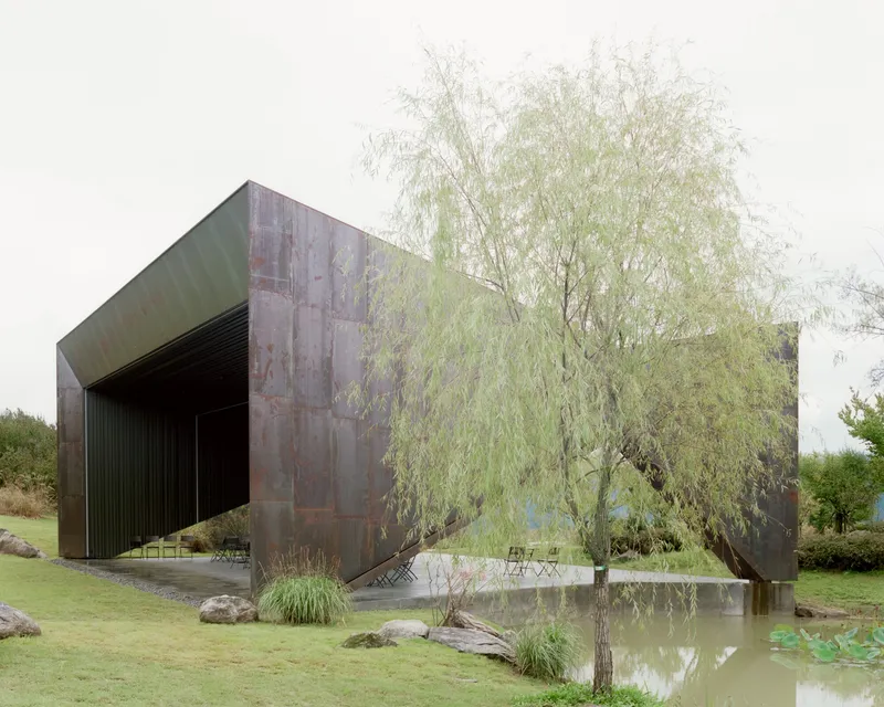 Corten steel exterior walls beside a shallow reflecting pool with willow tree and natural grasses