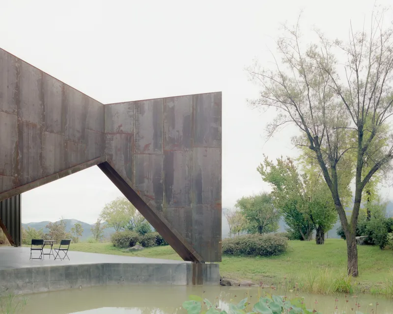 Angled steel support meeting the weathered panel facade above a concrete terrace and shallow pool