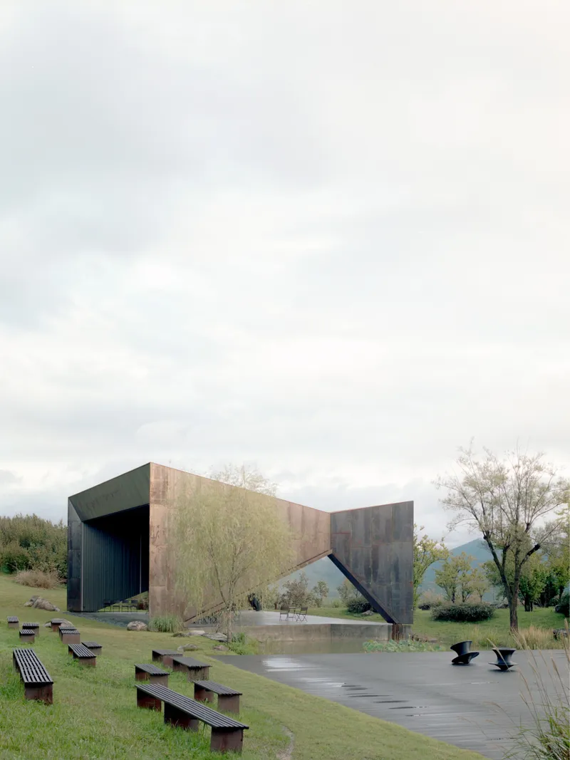 Side view of the folded steel structure with reflecting pool and benches under an overcast sky