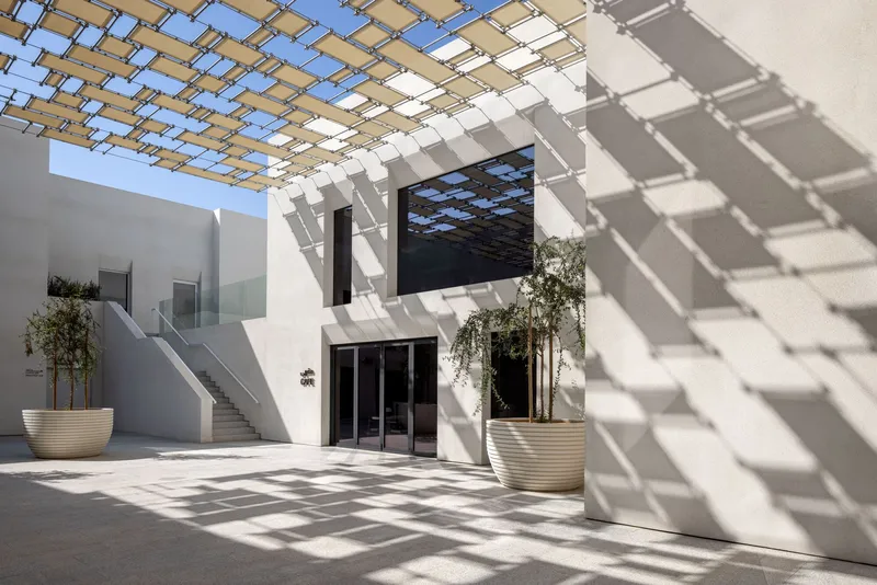 White courtyard with timber pergola casting geometric shadows across potted olive trees and staircase