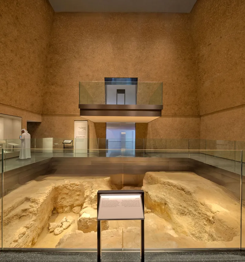 Museum gallery with excavated archaeological trench protected by glass balustrade under textured earthen walls