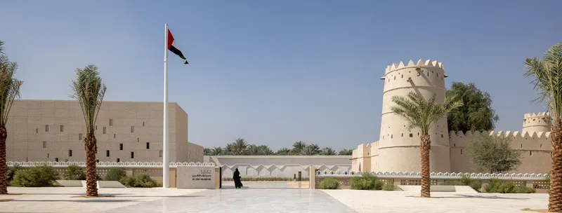 Plaza view with crenellated towers, white walls, palm trees and a flagpole under clear skies