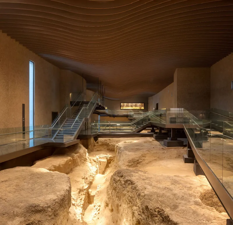 Interior exhibition space with glass walkways suspended over excavated archaeological remains beneath a curved timber ceiling