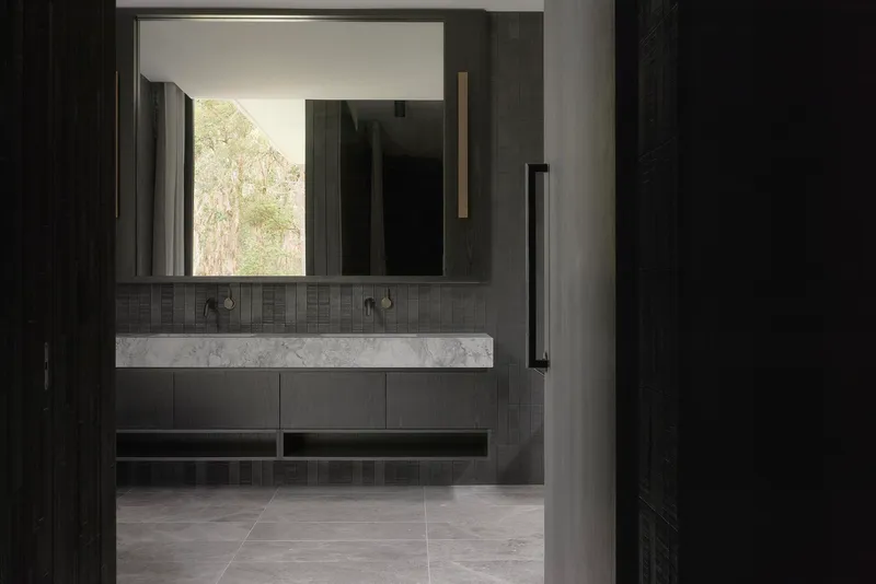 Bathroom vanity with marble countertop, dark tile and mirror reflecting trees outside