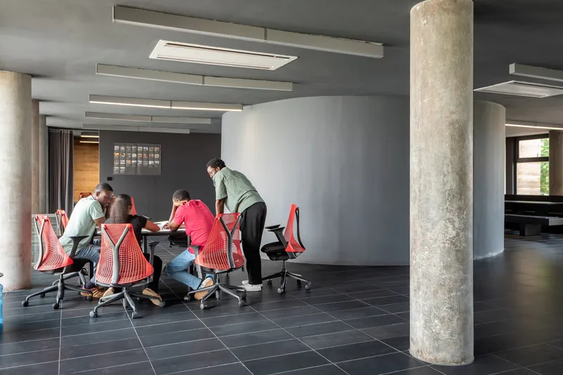 Collaborative workspace with exposed concrete columns, grey walls, and a group gathered around red mesh chairs